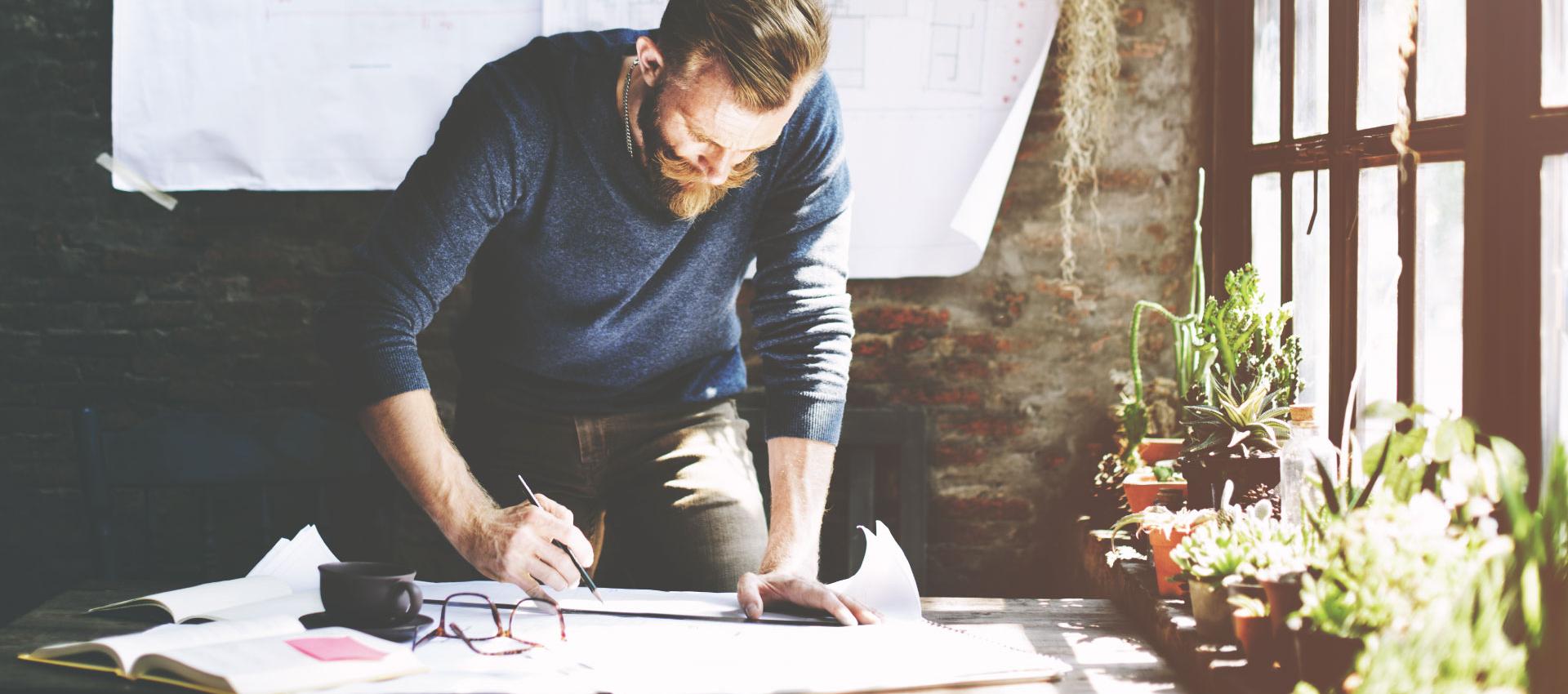 A man working on blue prints, with a cup of coffee and reading glasses on the table, with plants in the window.