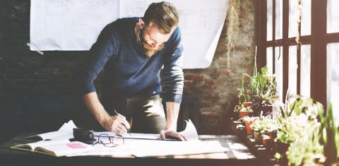 A man working on blue prints, with a cup of coffee and reading glasses on the table, with plants in the window.